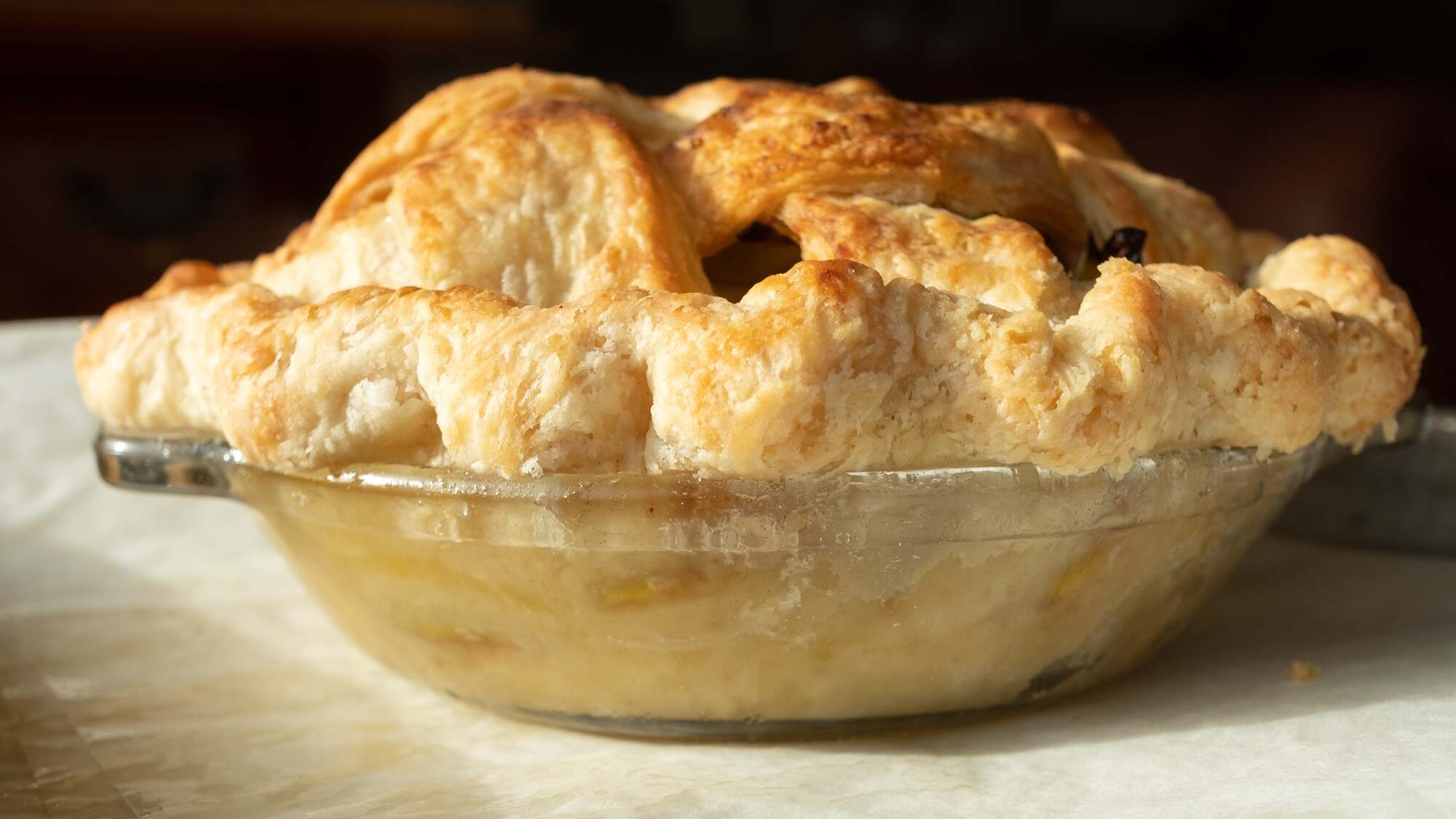 Side-facing photo of an apple pie with a lattice crust. It's in a glass plate, sitting on a parchment-lined baking sheet. The sides and bottom of the crust are so undercooked that you can see a few green-skinned apple slices poking through. 