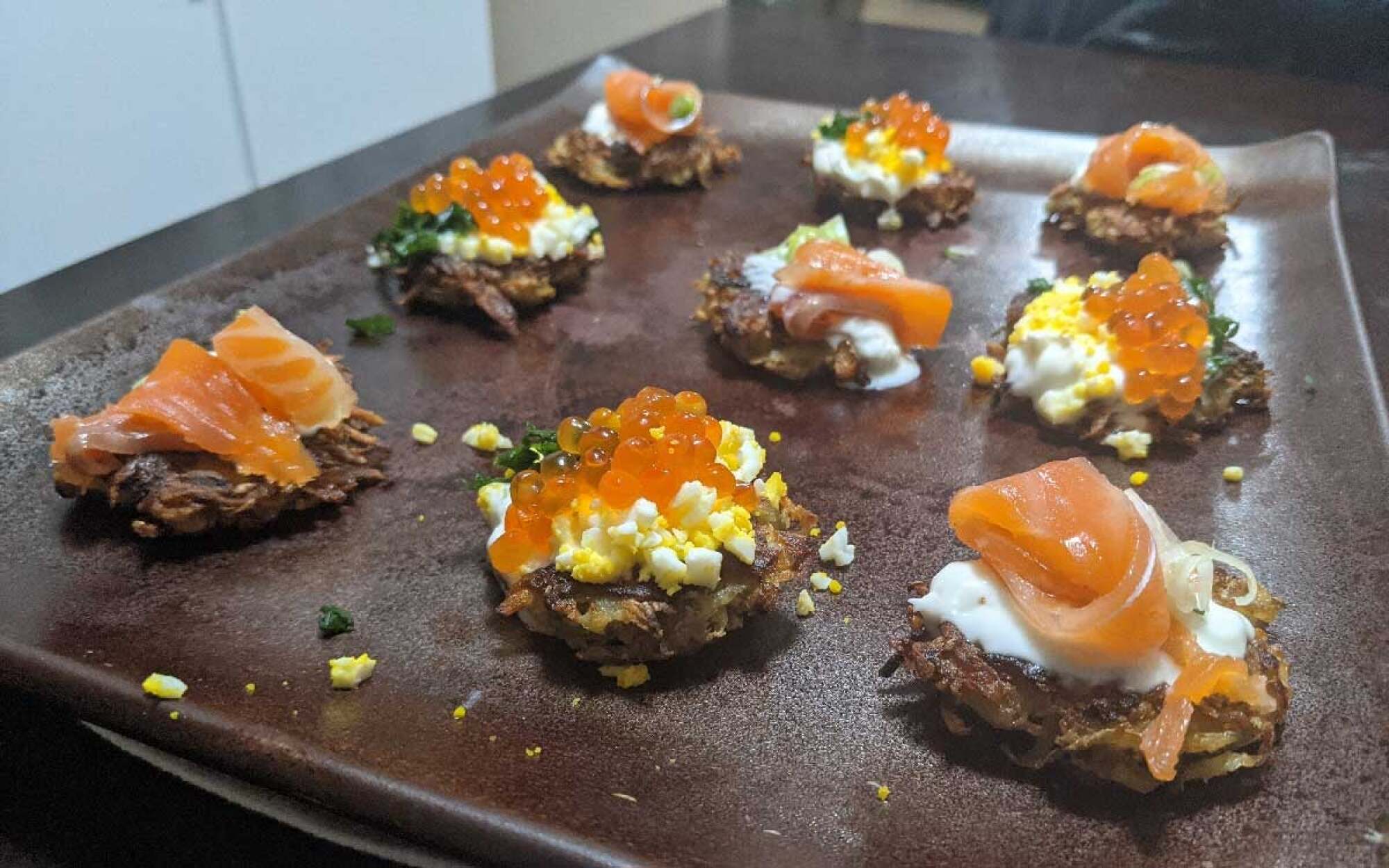 Latkes on a board with smoked salmon, hard boiled egg, row, chives, and creme fraiche. 