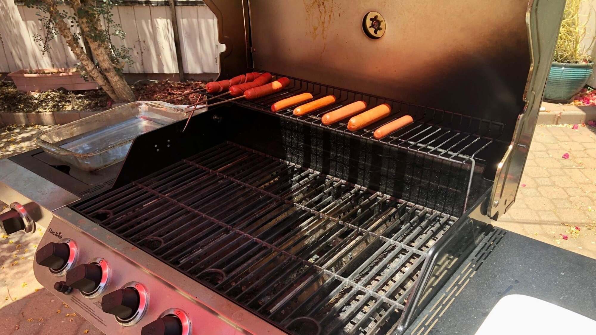 Multiple hot dogs sit on the top shelf of an open grill on a sunny day.