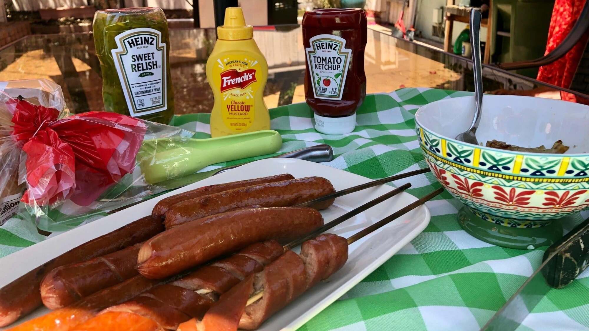 Grilled hot dogs stacked on a plate sit beside a table full of condiments and toppings.