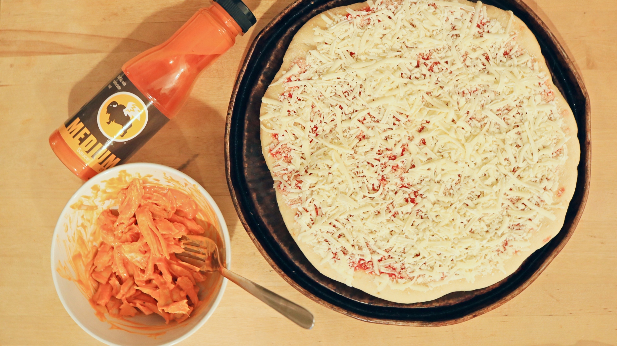 Pizza next to a bottle of buffalo sauce and a bowl of shredded meat.