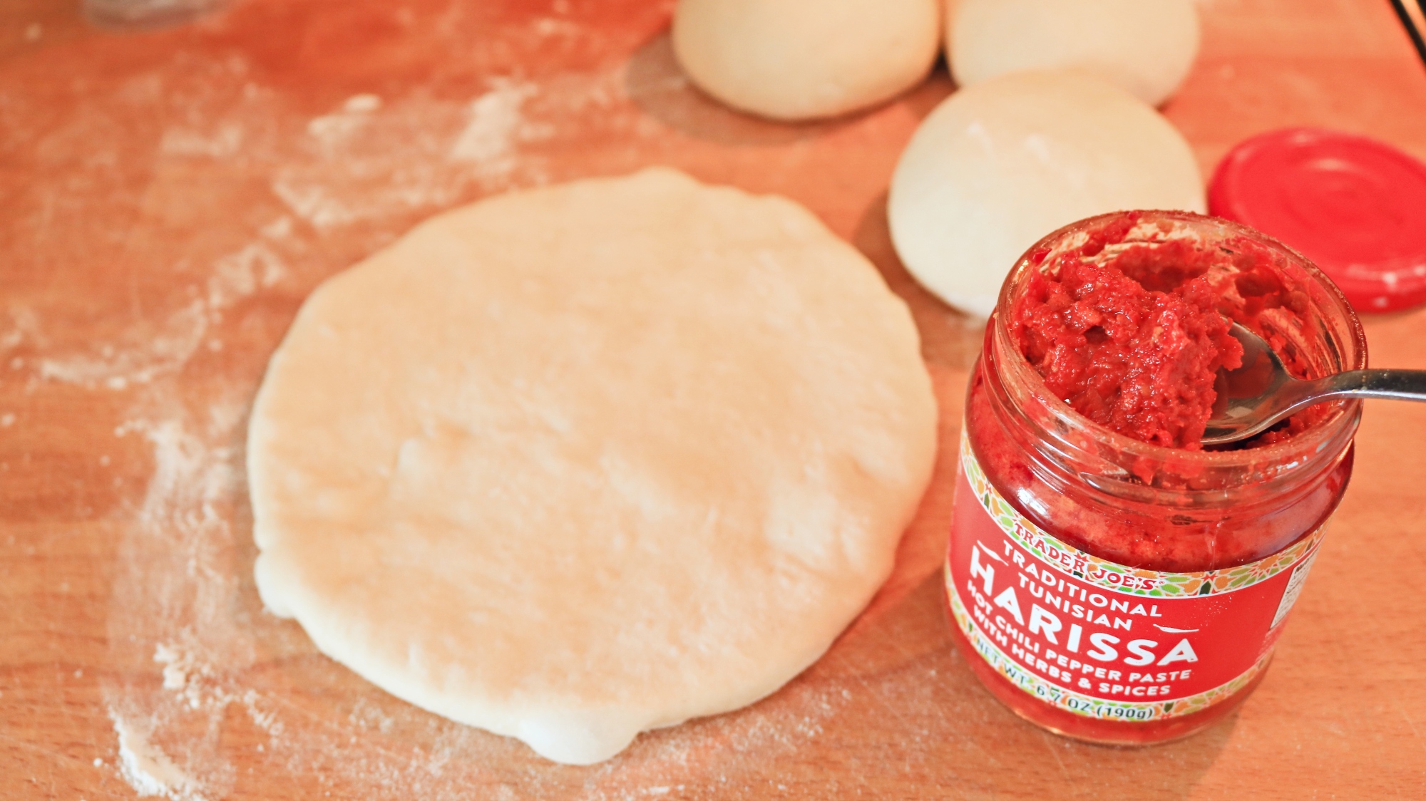 A jar of harissa sits next to a small round of pizza dough.